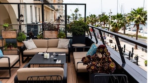 An outdoor seating area with a sofa, chairs, and tables, surrounded by potted plants and overlooking a waterfront with palm trees and boats. - Hôtel Restaurant Le Touring (Saint-Raphael)