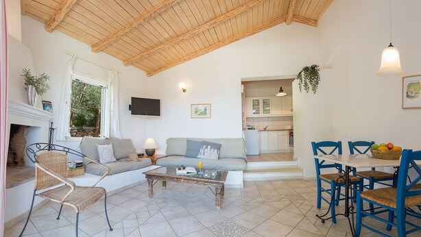 A cozy living room with a sofa, a coffee table, and wicker chairs. There is a kitchen in the background with a dining table and chairs.