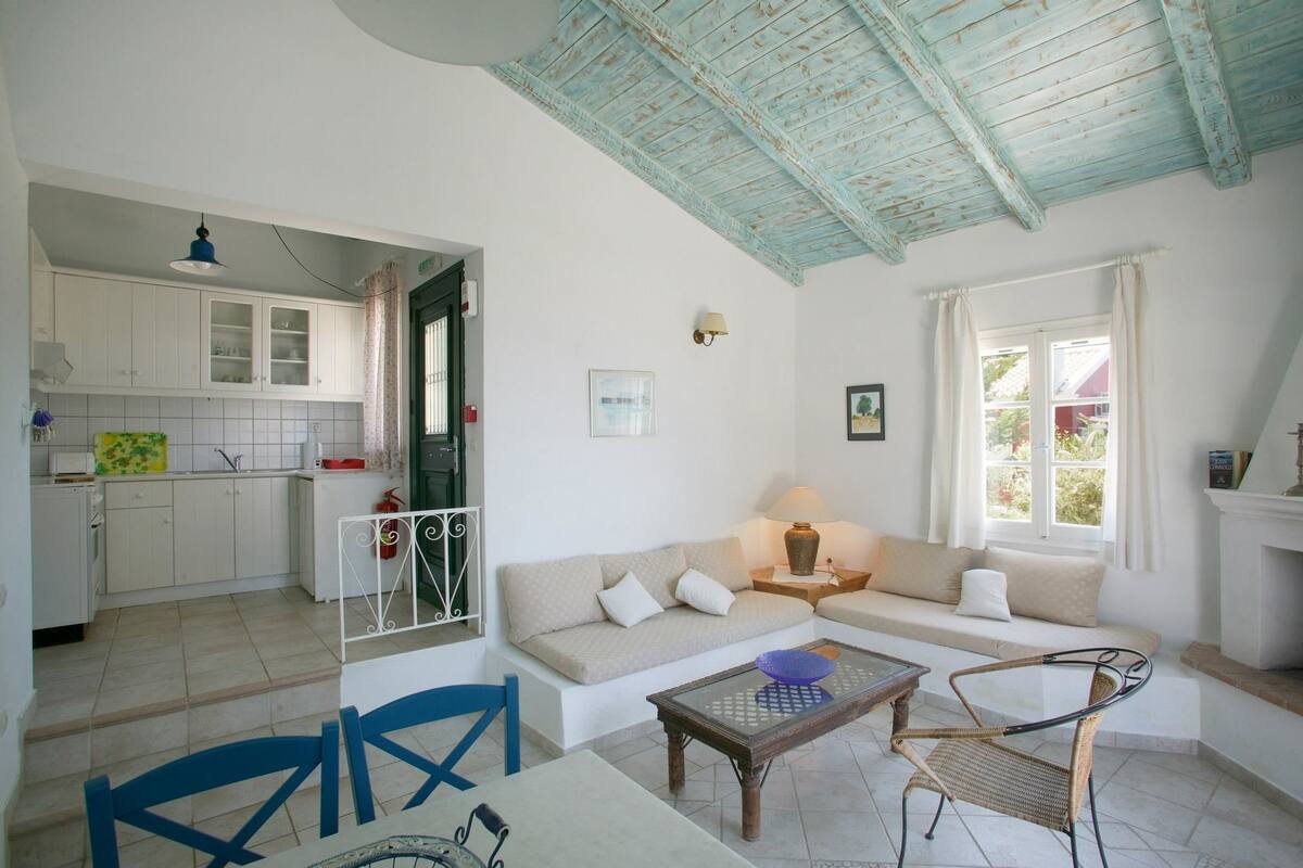 A living room with a sofa, a coffee table, and a dining area with chairs. There is a kitchen in the background with cabinets and a window.