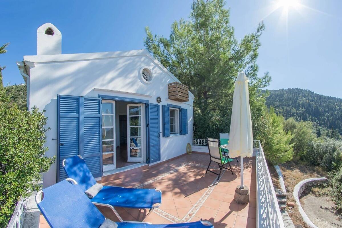 A white building with blue shutters, a patio with a table and chairs, and a view of a forested hill.
