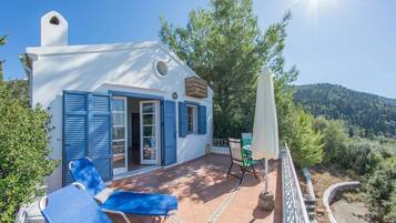 A white building with blue shutters, a patio with a table and chairs, and a view of a forested hill.
