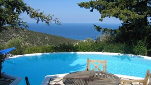 A pool with a wooden table and chairs, surrounded by greenery and overlooking a body of water.