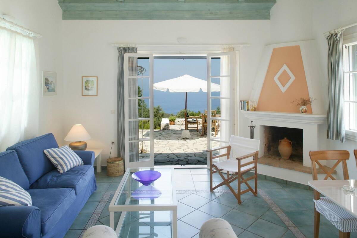 A living room with a blue sofa, a glass coffee table, and a fireplace. There is a view of the outdoors through open doors.