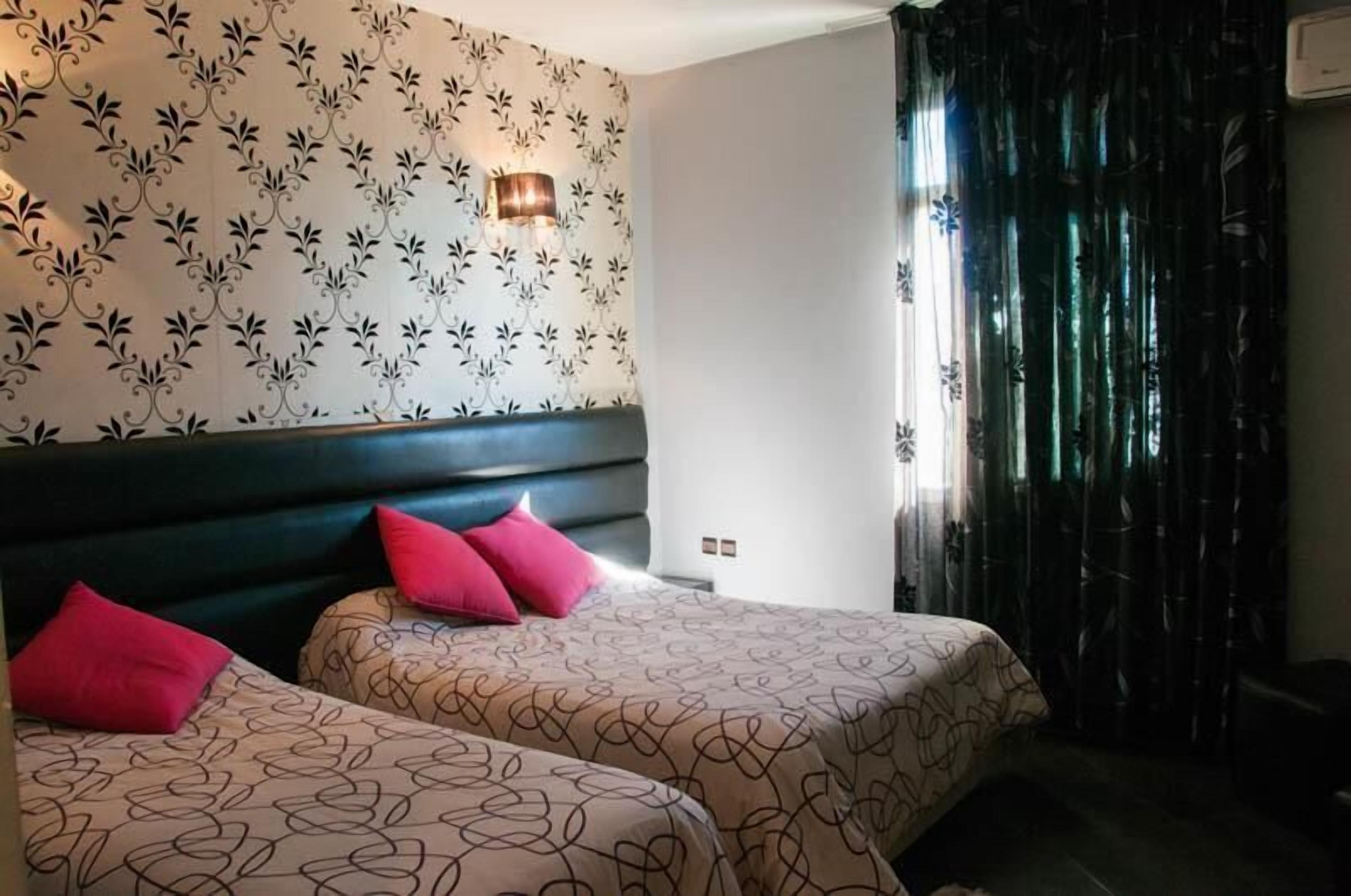 A hotel room with two beds, a patterned wall, a wall-mounted lamp, and a window with curtains.