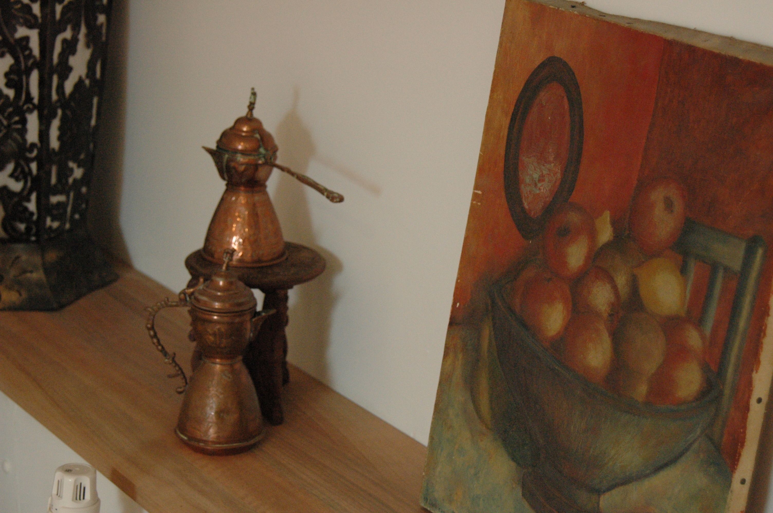 Een koperen theepot en suikerpotje op een houten plank, naast een schilderij van appels in een mand.