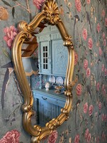 A room with a large ornate mirror, floral wallpaper, and a blue dresser with decorative plates.