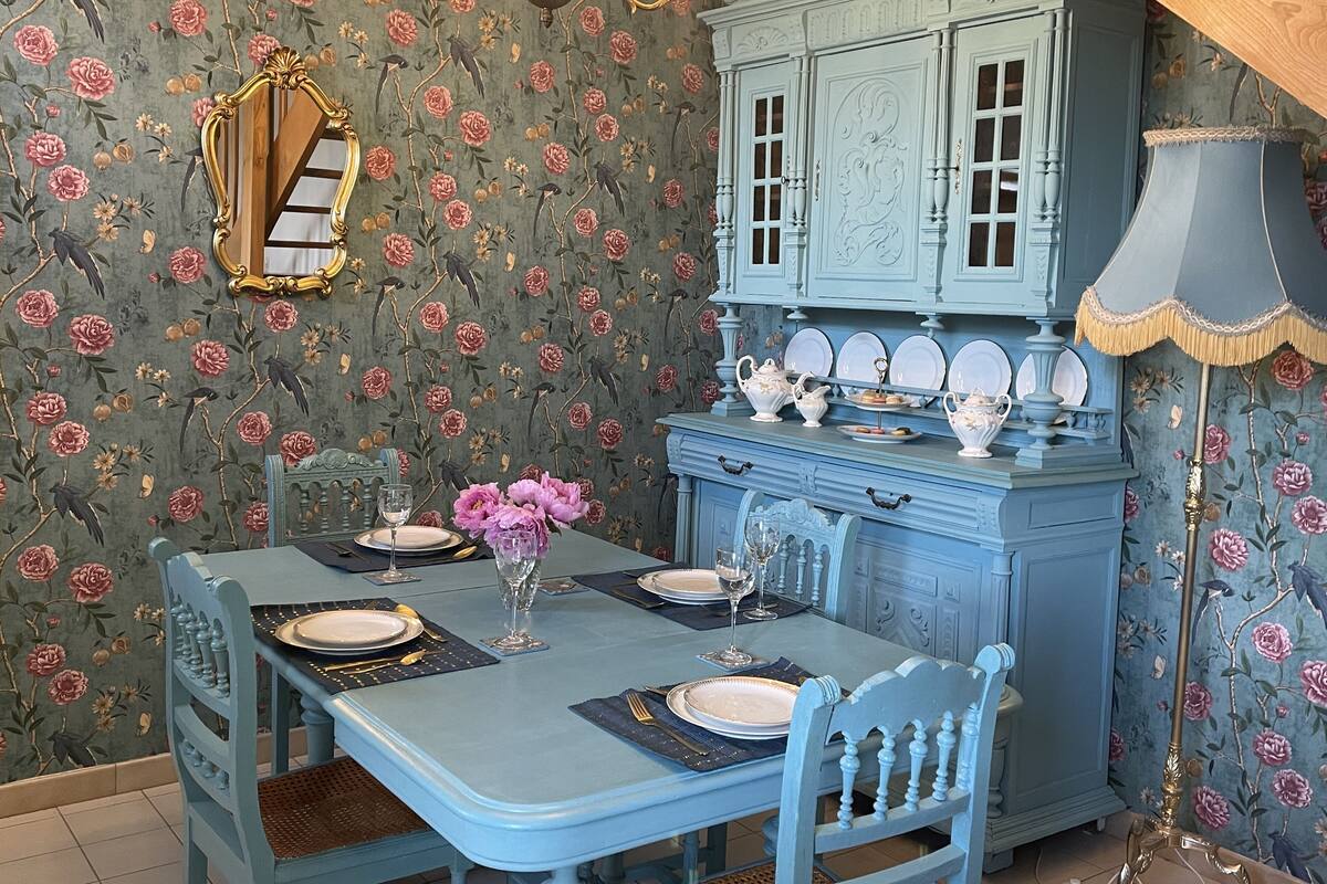 Un coin repas avec une table bleue dressée pour quatre personnes, un papier peint à motifs floraux, un buffet bleu d’époque et un lustre.