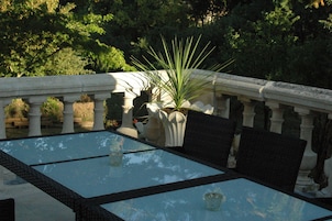 A glass-top table with wicker chairs, surrounded by a stone balustrade and lush greenery.