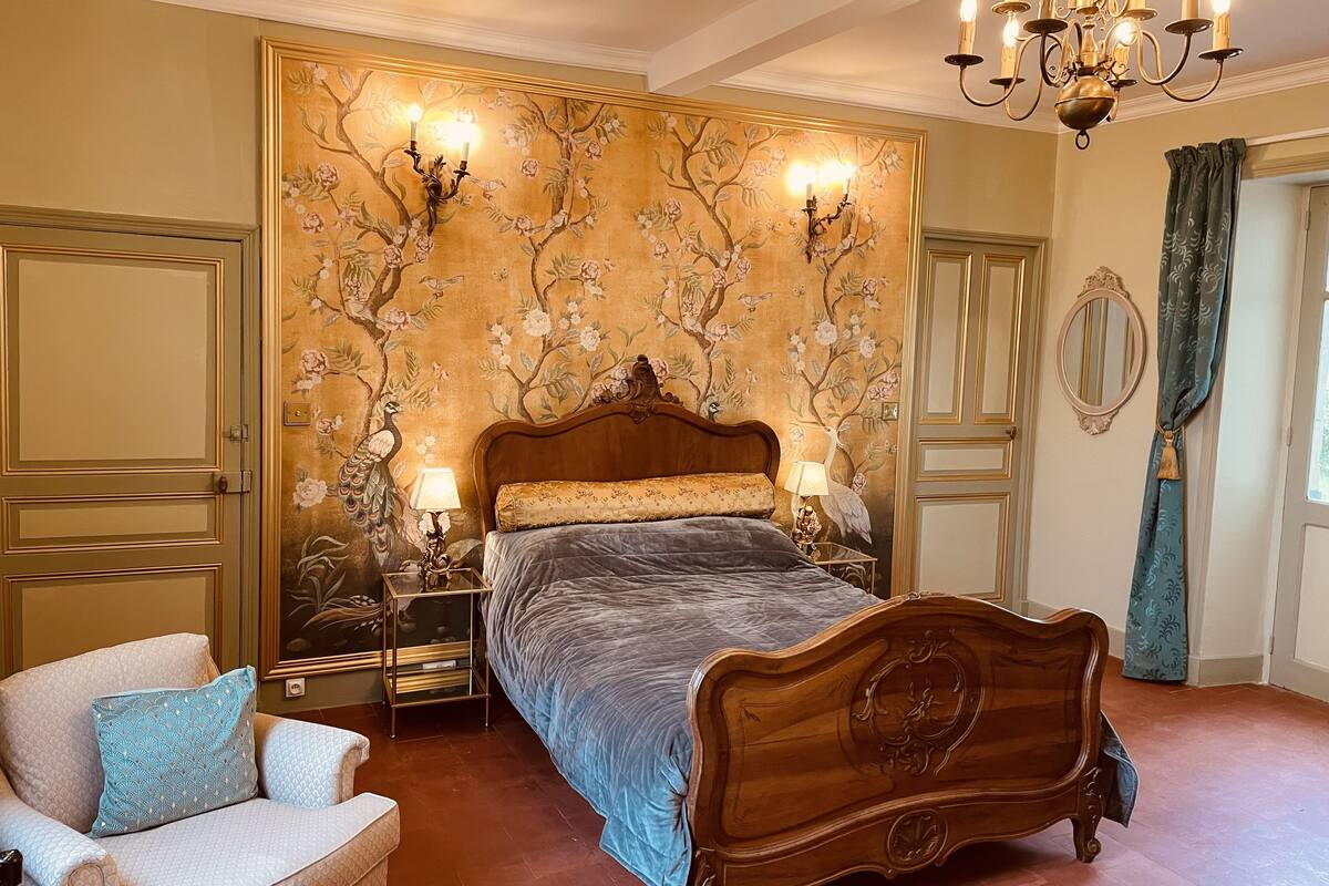 Une chambre à coucher avec un grand lit, deux tables de chevet, un lustre, une chaise et un papier peint à motifs floraux.