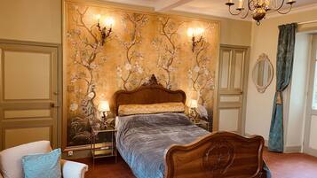 A bedroom with a large bed, two bedside tables, a chandelier, a chair, and a floral wallpaper design.
