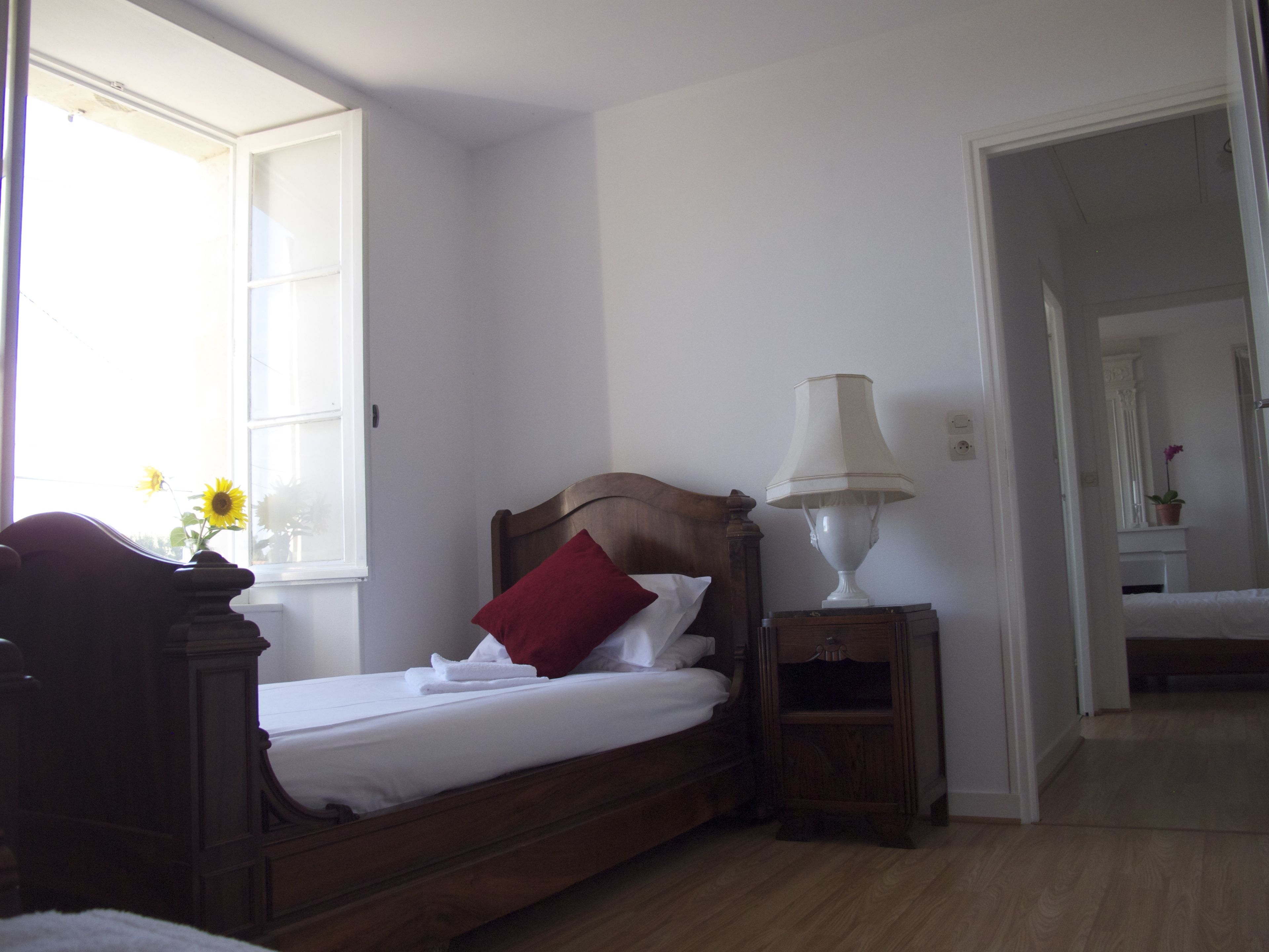 Une chambre à coucher avec un lit en bois, du linge de lit blanc, un oreiller rouge, une lampe de chevet et une fenêtre donnant sur un tournesol.