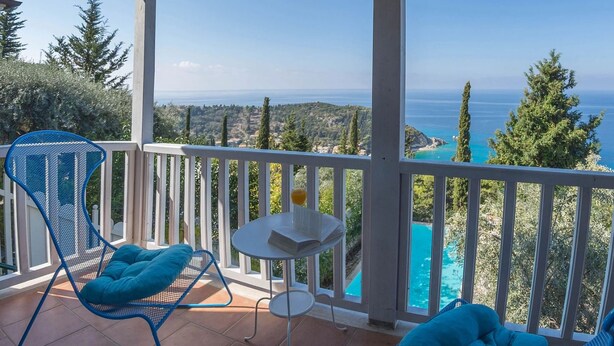 Un balcon avec deux chaises bleues et une petite table, donnant sur une piscine turquoise et un paysage pittoresque.