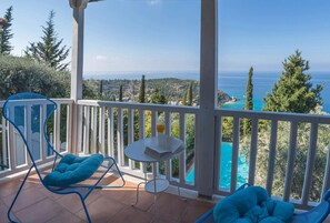 Un balcon avec deux chaises bleues et une petite table, donnant sur une piscine turquoise et un paysage pittoresque.