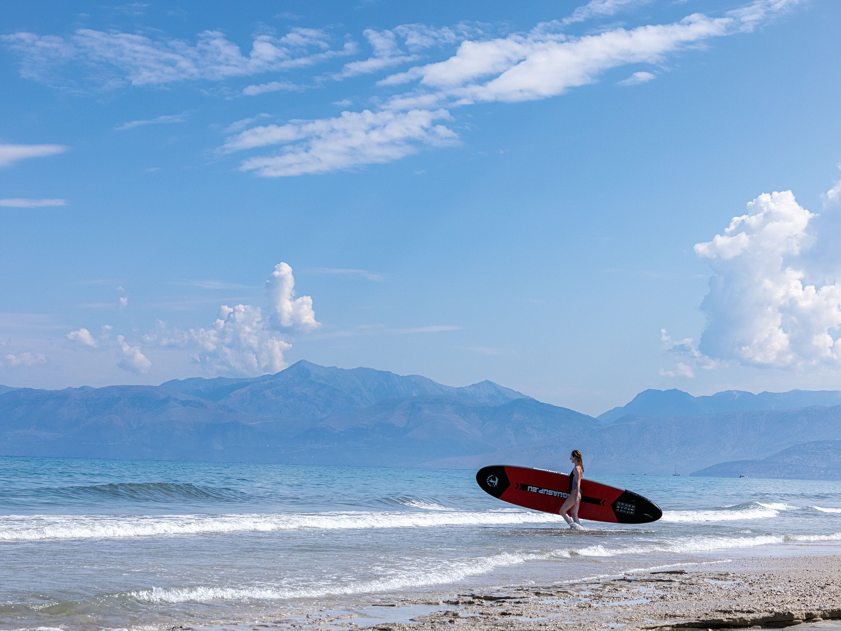 Ein Strand mit Bergen im Hintergrund und einer Person, die einen Surfbrett trägt.