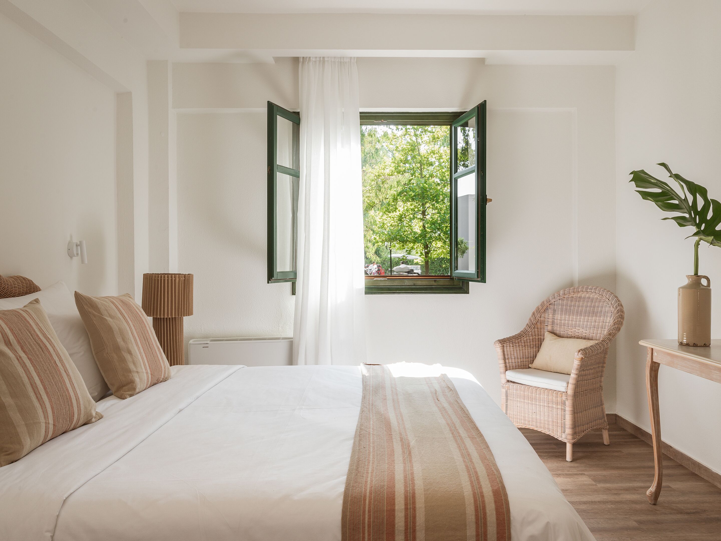 Un dormitorio con cama, una silla, una mesa y ventana con vista a follaje.