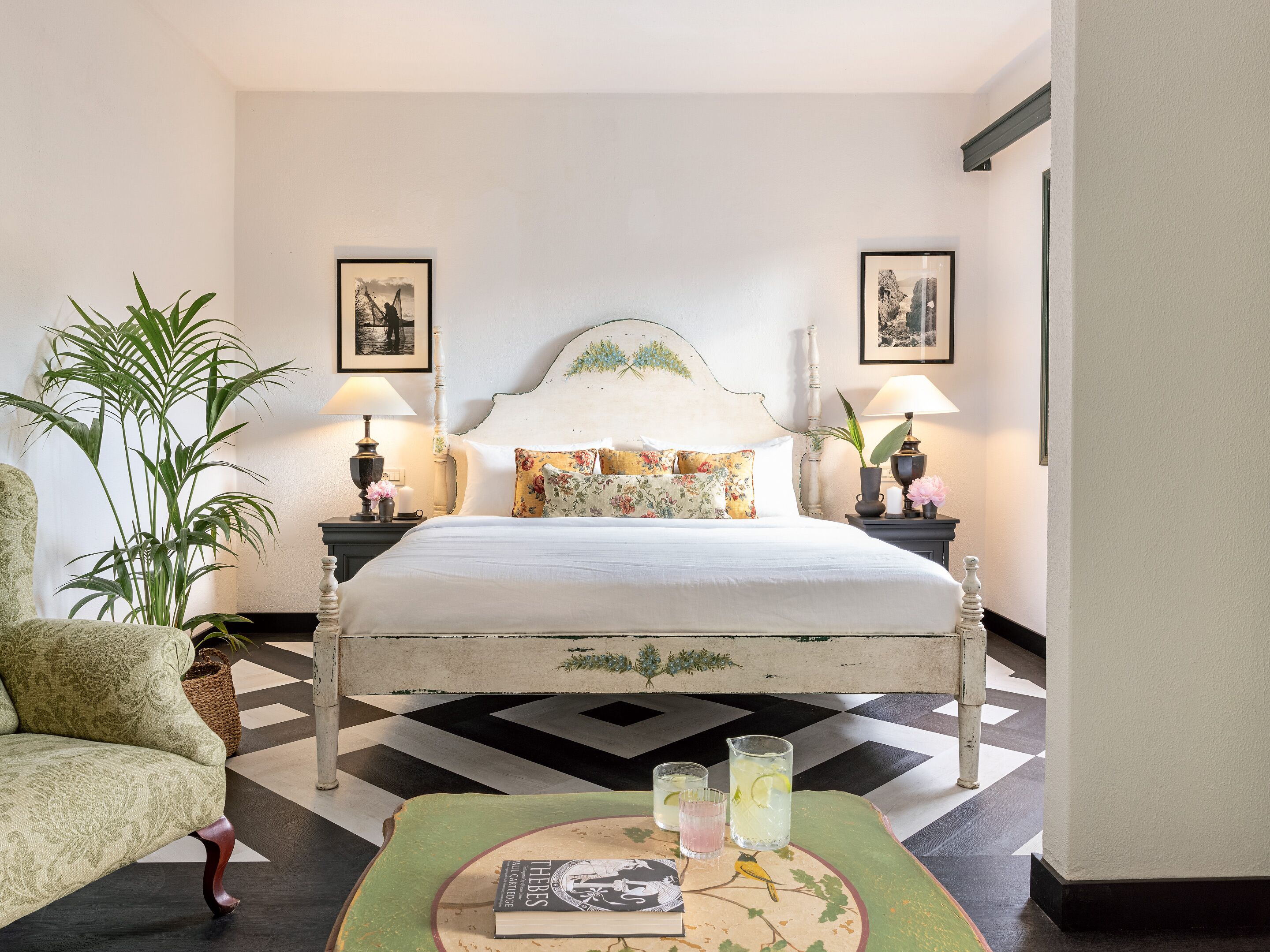 Un dormitorio de hotel con una cama, dos mesitas de noche, una silla, una planta en maceta y una mesa con un libro y vasos.