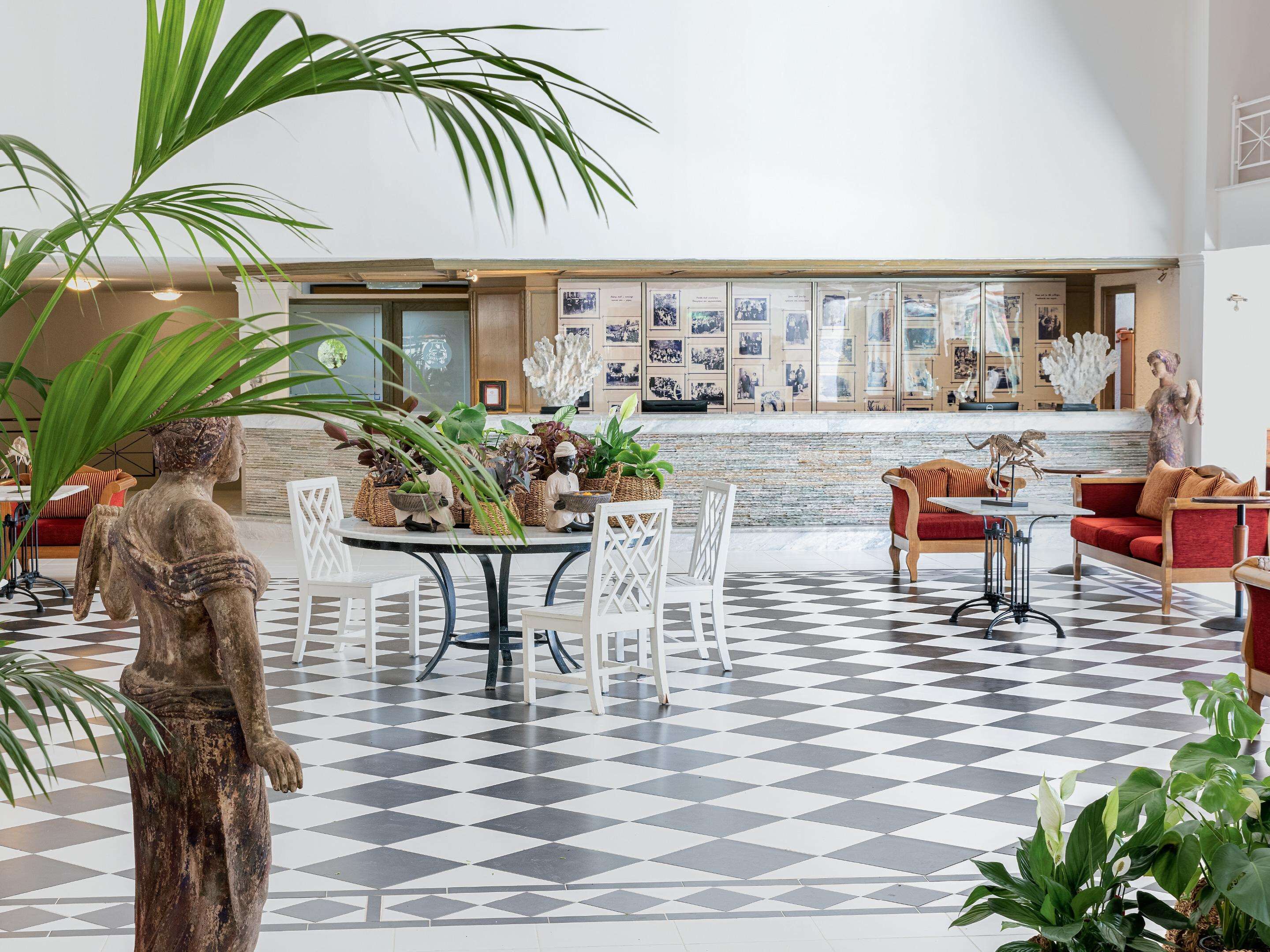 Un hall d’entrée spacieux avec un sol à carreaux, un espace de restauration central avec des chaises blanches et un canapé rouge.