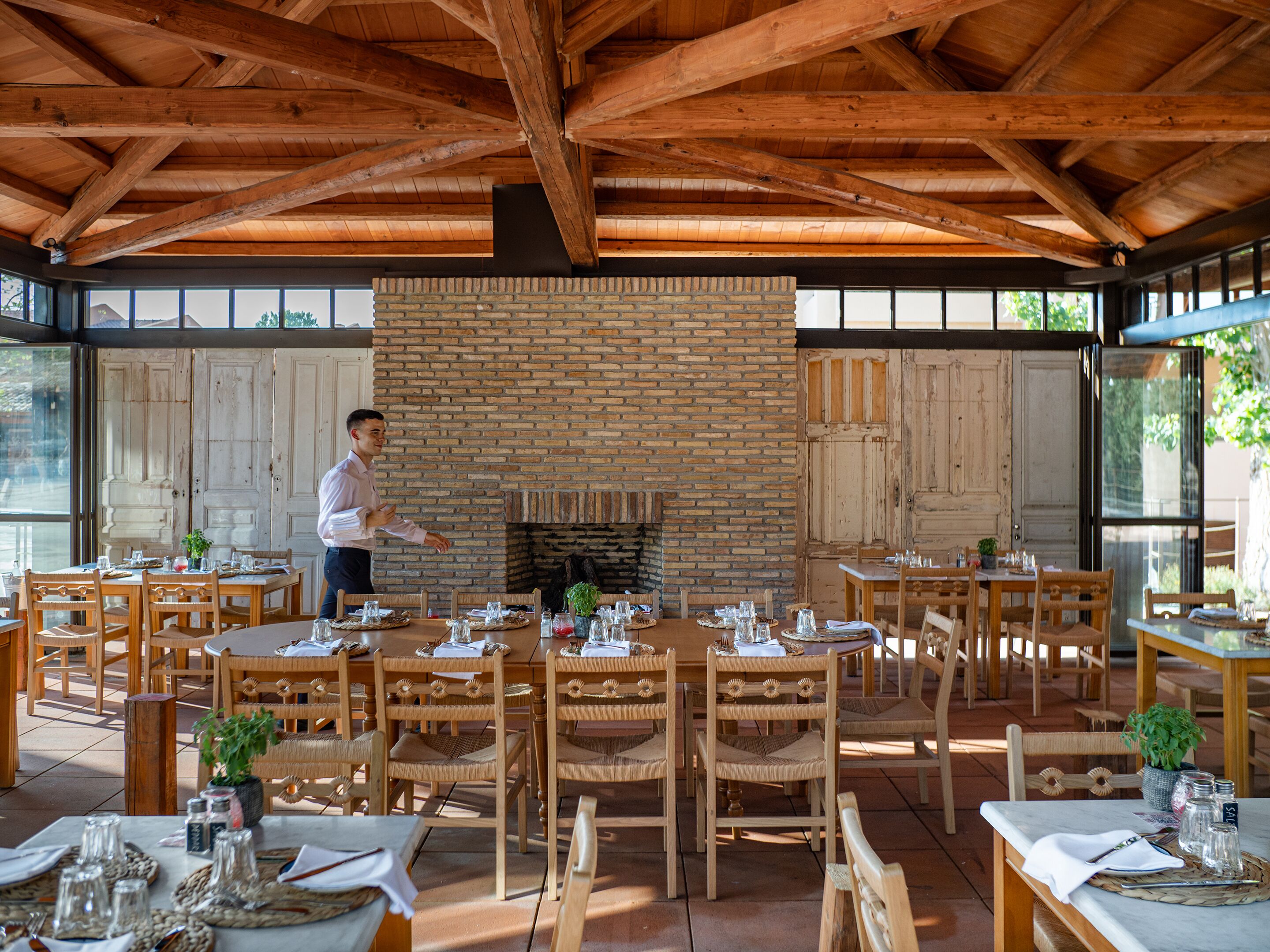 A restaurant with a brick fireplace, wooden tables and chairs, and a person serving.