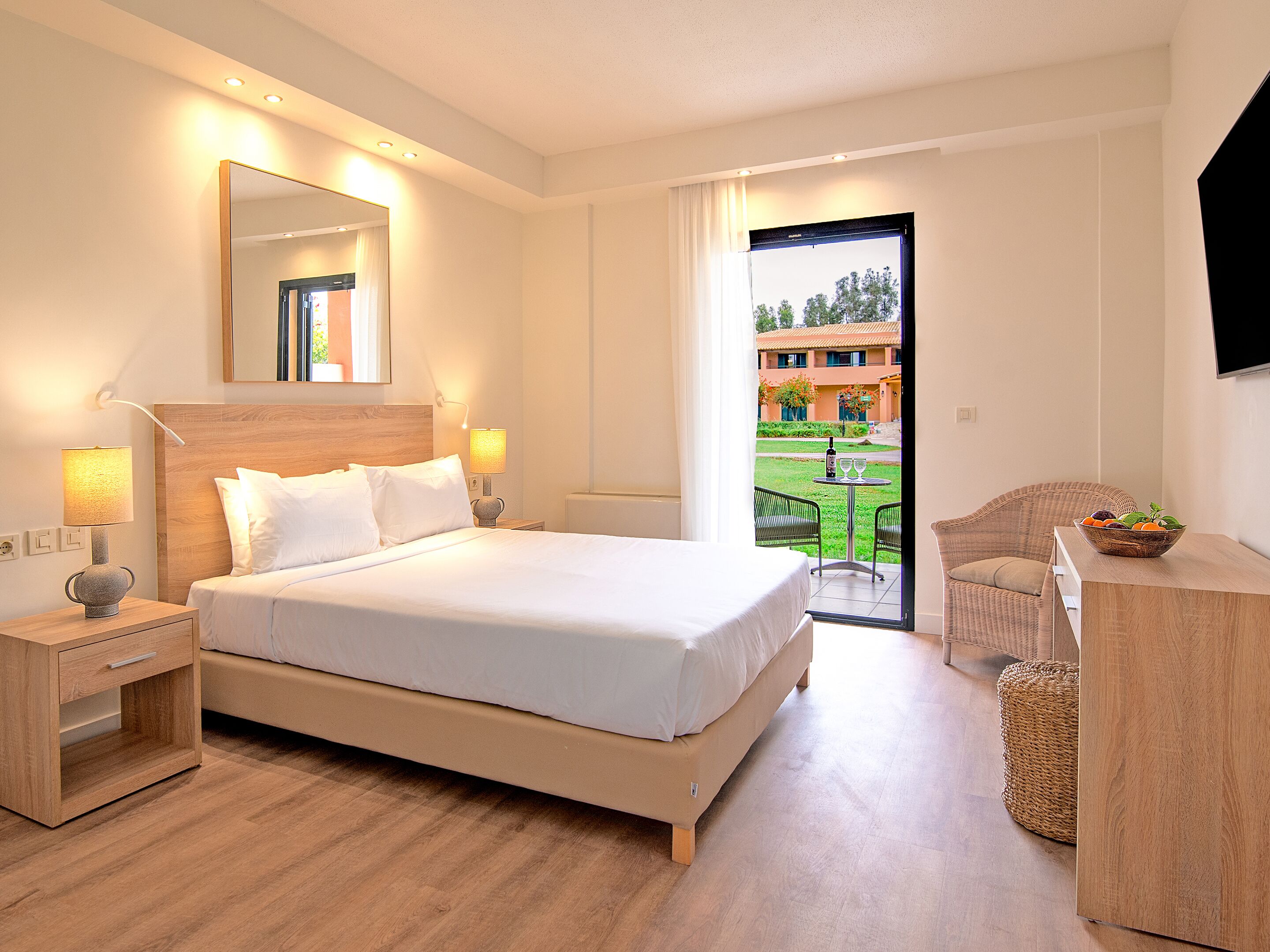 Habitación de hotel con una cama grande, mesitas de noche, un espejo, un televisor y vistas al jardín exterior a través de la puerta.
