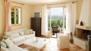 Un salón amplio con un sofá blanco, una mesa de centro y una chimenea. Se ve follaje verde a través de las ventanas.