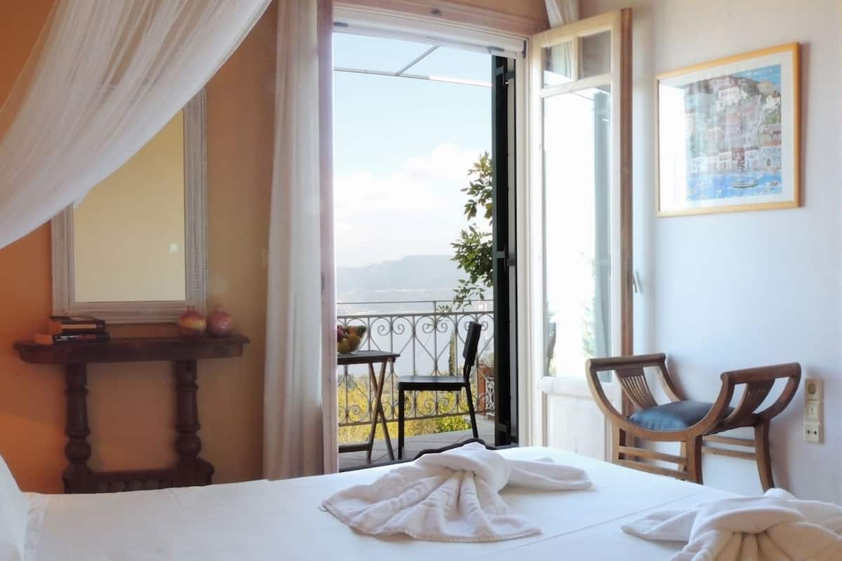 A hotel room with a bed, a table with a lamp, a chair, and a balcony with a view.