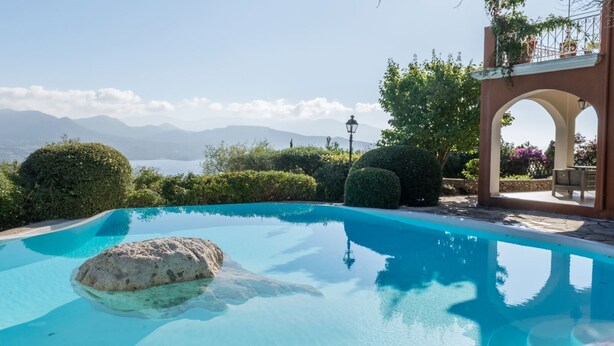 Una piscina azul cristalina con una roca en el centro, rodeada de arbustos cuidados y una vista de montañas al fondo.