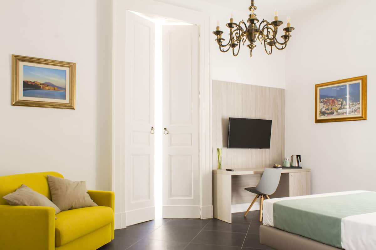 Un sillón amarillo brillante con un cojín, una cama con colcha verde, un escritorio con una silla, un televisor de pantalla plana montado en la pared y una araña.