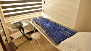 A small, single-bed room with a massage chair, a control panel, and a window with blinds.