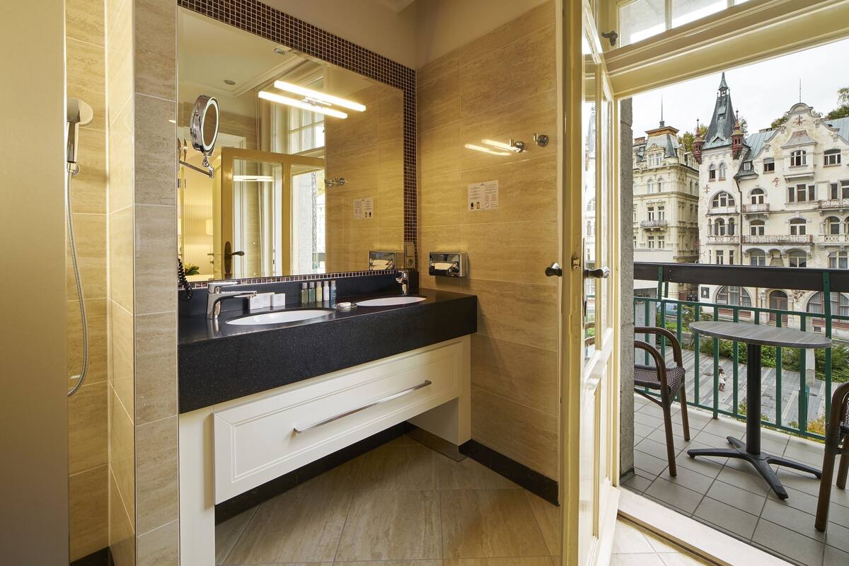 Casa de banho com um espelho grande, duas pias e vista para um edifício histórico através de uma porta de vidro.