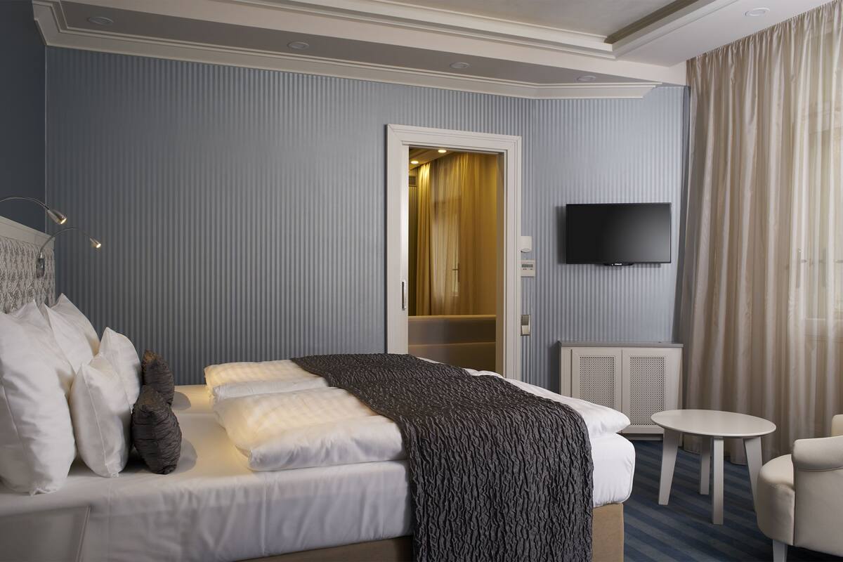 Quarto de hotel com uma cama, uma mesinha, uma cadeira, uma televisão e uma porta que dá para um banheiro.
