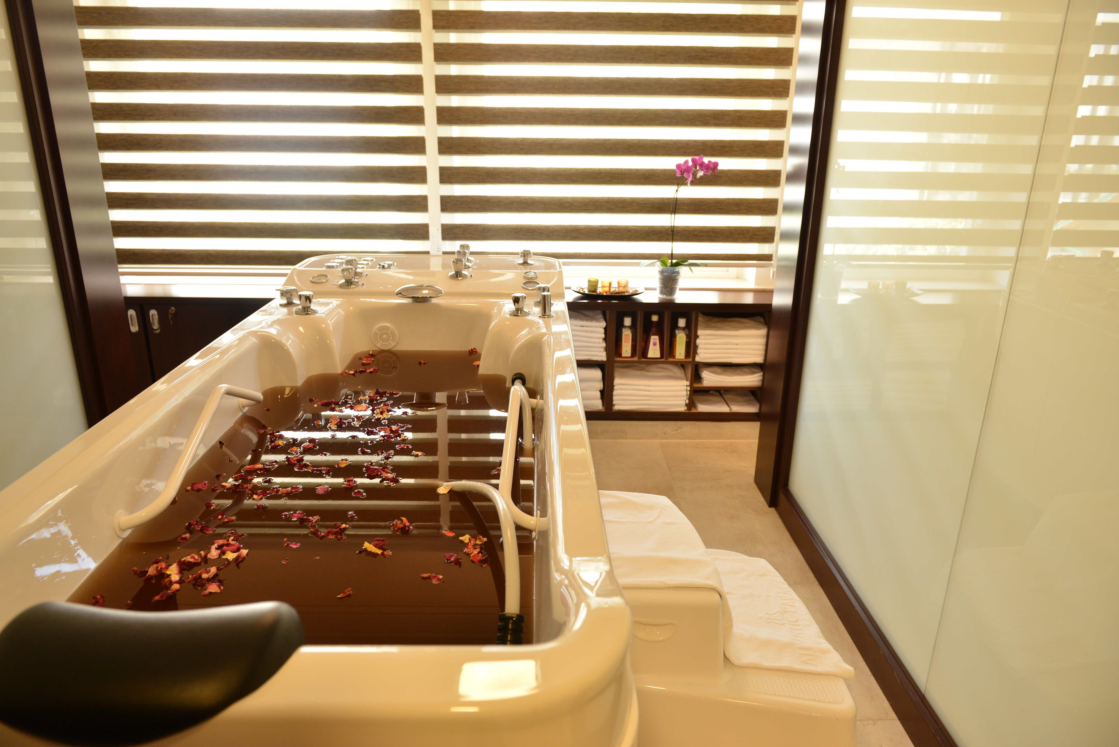 A spa bath with rose petals, a wooden bench, and a table with towels and a vase of flowers.