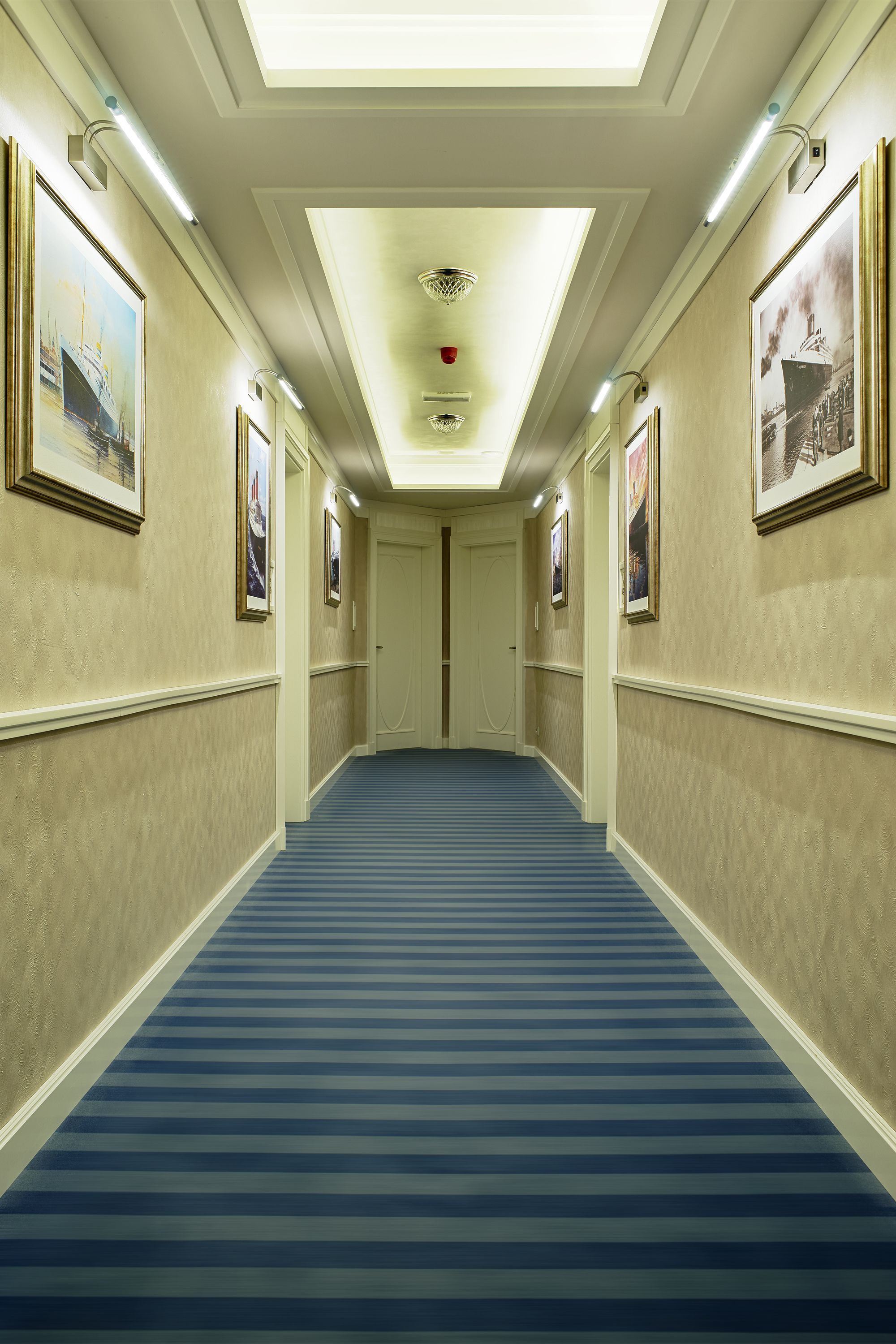 Un pasillo de hotel con alfombra azul, cuadros en las paredes y un candelabro en el techo.