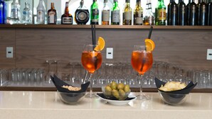 Zona de bar con dos bebidas, cuencos de aceitunas y patatas fritas, y una variedad de botellas de licores al fondo.