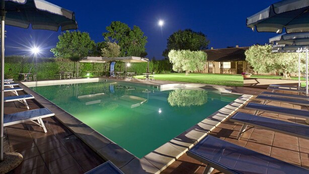 Piscina com espreguiçadeiras e guarda-sóis, rodeada por árvores e um terraço bem iluminado.