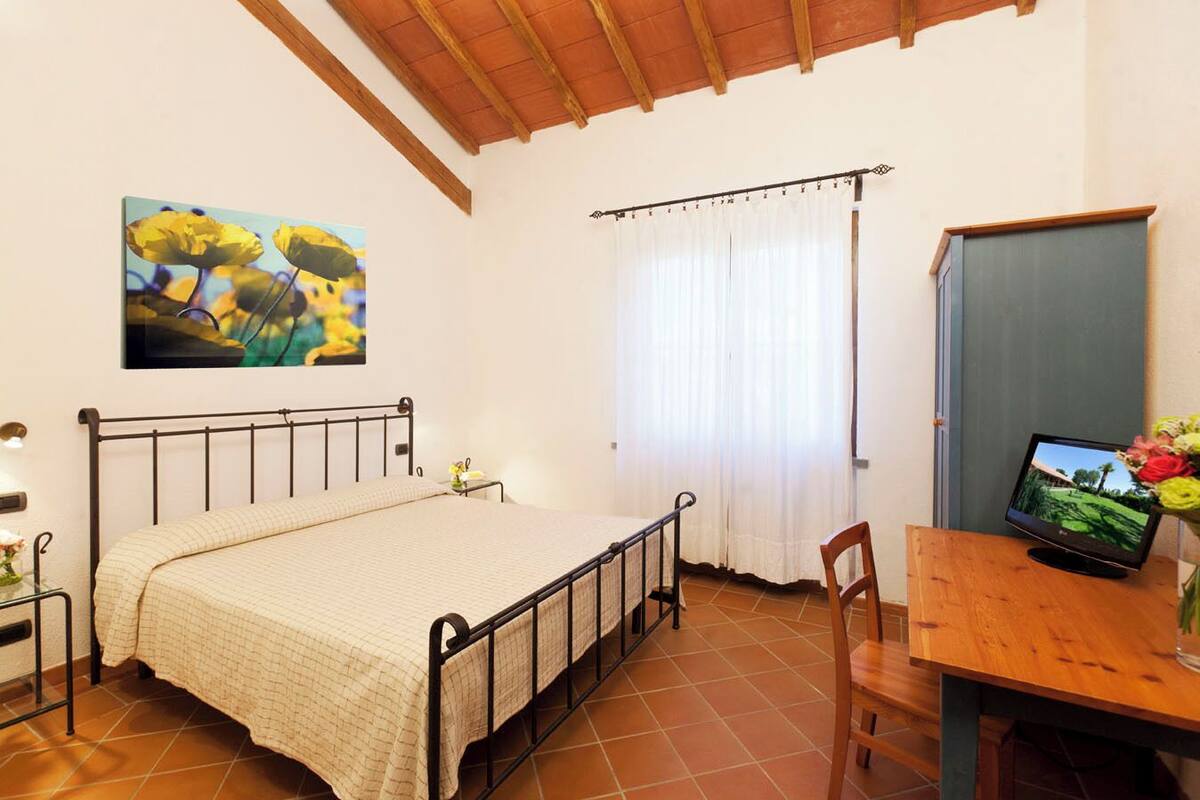 Een slaapkamer met een bed, een bureau met een computer, een stoel, een raam met gordijnen en een schilderij aan de muur.