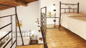 Quarto com piso de madeira, cama com colcha branca, estrutura de cama em metal preto, mesinha de cabeceira com um vaso de flores e escada com corrimão de metal.