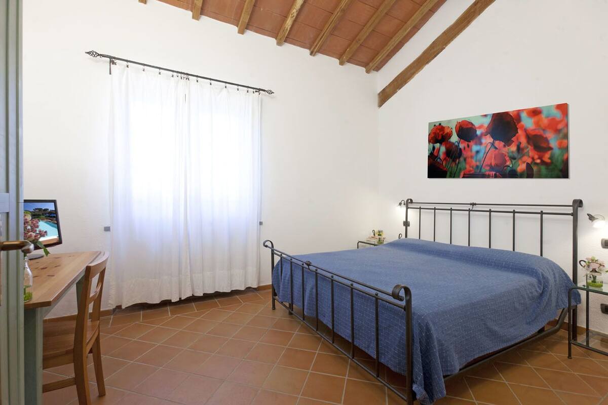 Een slaapkamer met een bed, een bureau, een stoel, een raam met gordijnen, een schilderij en een televisie op een standaard.