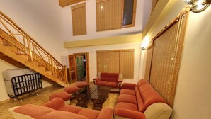 A spacious living room with a high ceiling, a staircase, and a view of the outside through large windows with blinds. - Daffodil Cottages (Manali)
