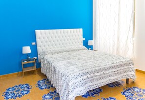 A bed with a white headboard and footboard, flanked by two bedside tables with lamps, set against a blue wall and a patterned floor. - B&B Le Marinelle (Salerno)