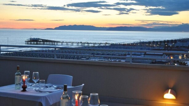 A dining area with tables set for two, overlooking a coastal view with a pier and mountains in the distance. - Hotel Capri & Residence (Camaiore)