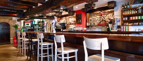 A bar area with wooden chairs, a counter with various bottles, and a stone wall in the background.