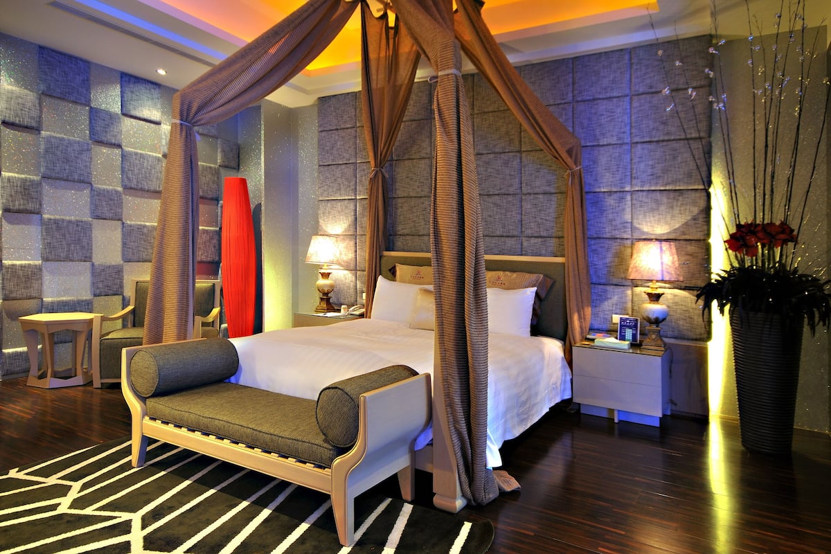A four-poster canopy bed with a nightstand and lamp, a chaise lounge, and a decorative vase with red flowers.