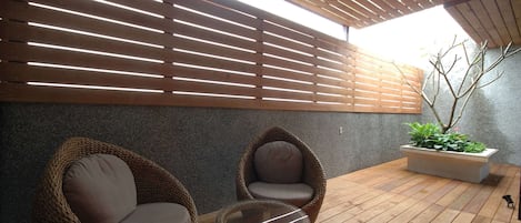 A covered outdoor seating area with two wicker chairs and a glass table, a potted plant, and a wooden floor.