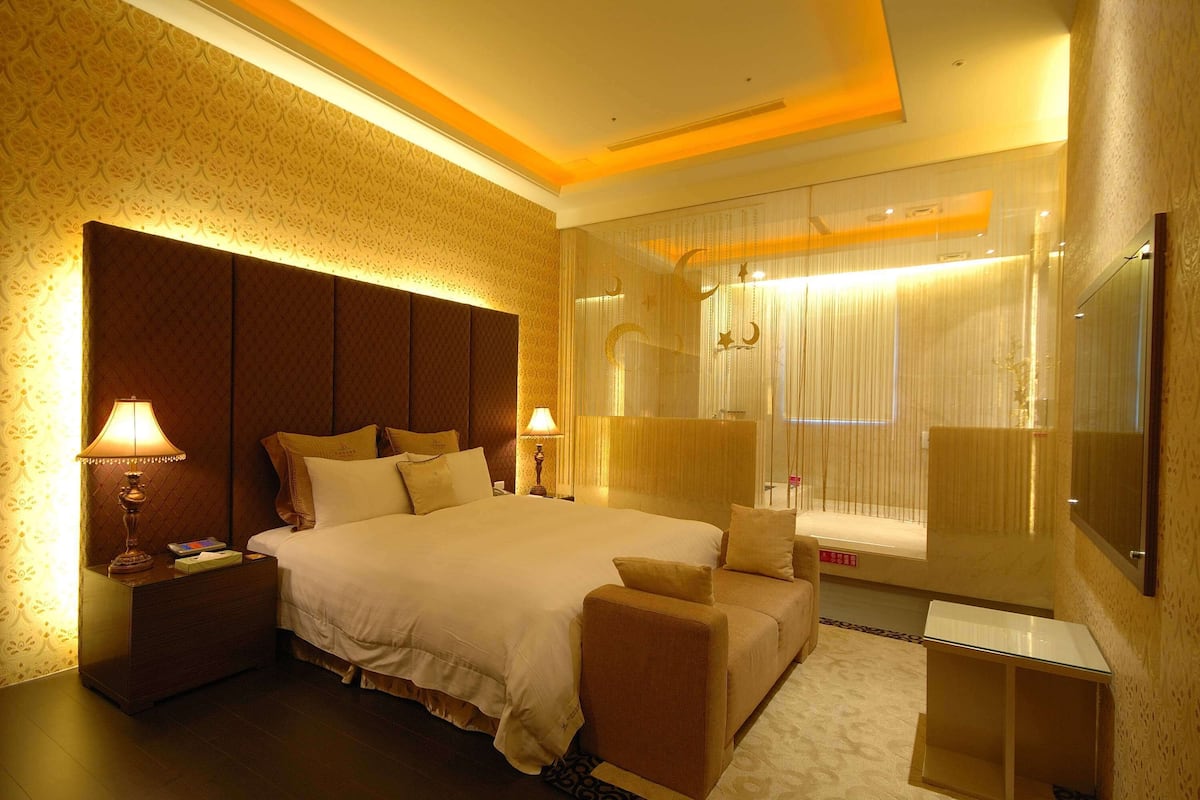 A hotel room with a large bed, two bedside tables with lamps, a sofa, a TV, and a window with curtains.