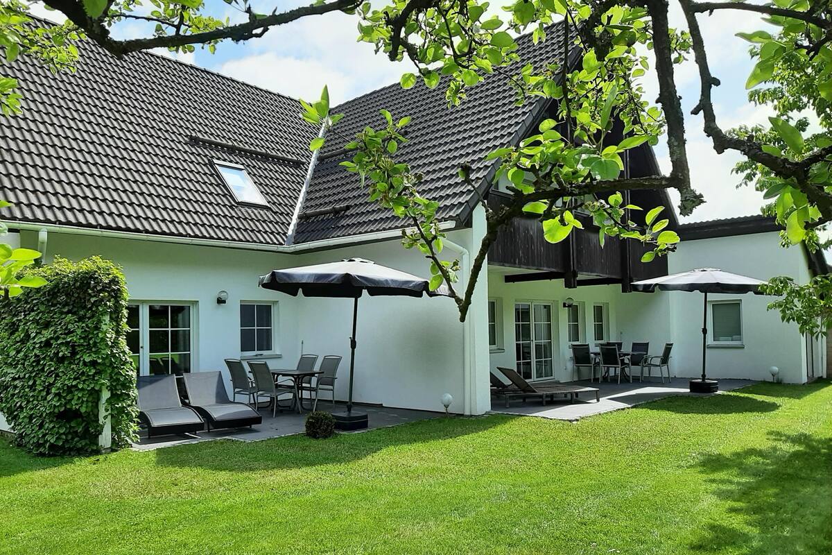 Une maison à deux étages, avec un toit en tuiles, un patio couvert aménagé avec du mobilier d’extérieur et une pelouse bien entretenue.