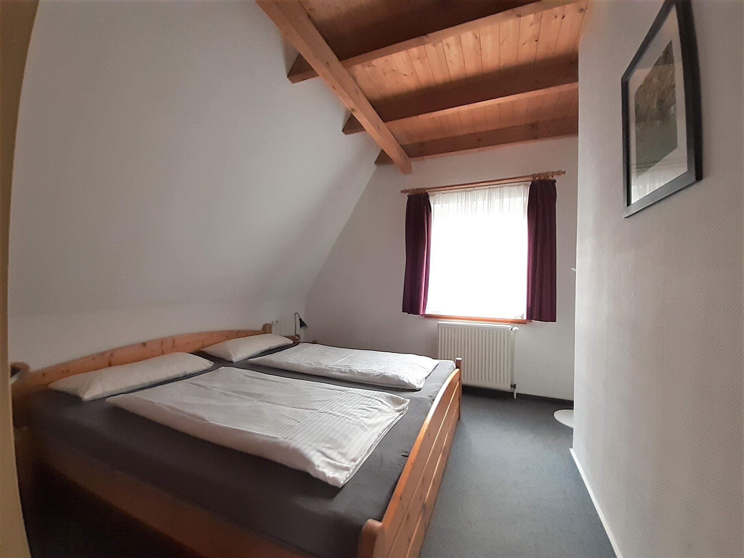 A small, single-room bedroom with a sloped ceiling, a window with curtains, and a radiator.