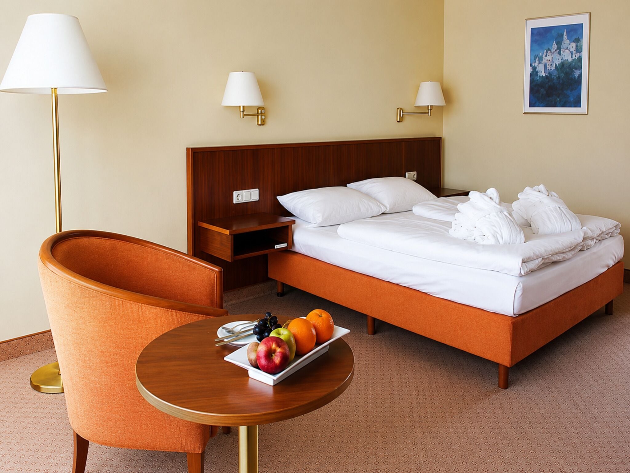 Habitación de hotel con cama, mesitas de noche, una silla, una pequeña mesa con fruta y lámparas de pared.