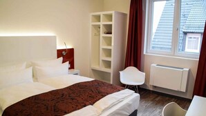 Habitación de hotel con cama, armario blanco, silla blanca y ventana con vistas a edificios.