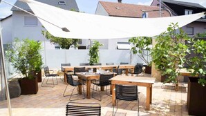 Un patio al aire libre con varias mesas y sillas, rodeado de plantas en macetas y un toldo blanco.
