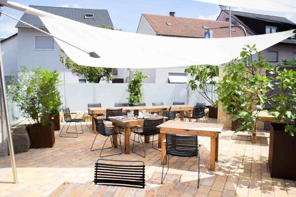 Un patio al aire libre con varias mesas y sillas, rodeado de plantas en macetas y un toldo blanco.